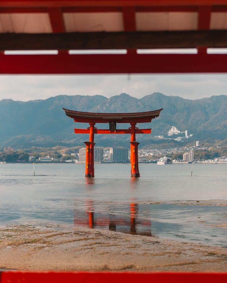 A traditional red torii gate stands in the water, framed by a wooden structure in the foreground. In the background, a cityscape and green mountains are visible under a cloudy sky.