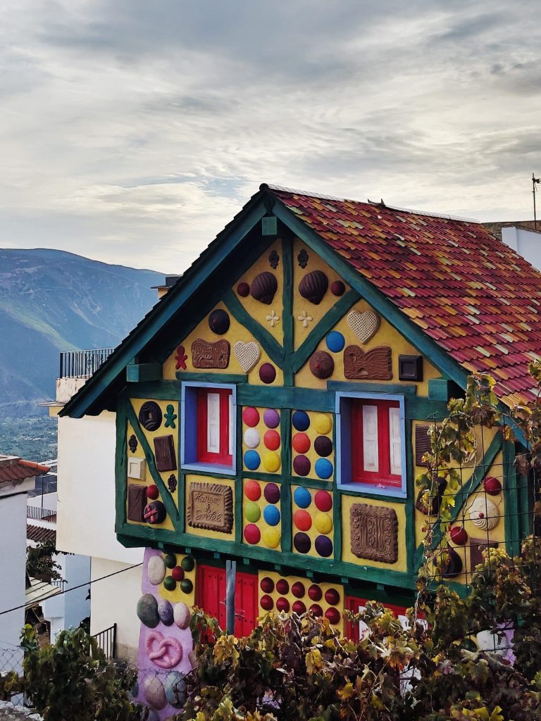 A colorful house with red, yellow, and green accents, decorated like a gingerbread house with candy designs, sits under a cloudy sky with Alpujarra mountains— Soportujar is one of the best day trips from Granada.