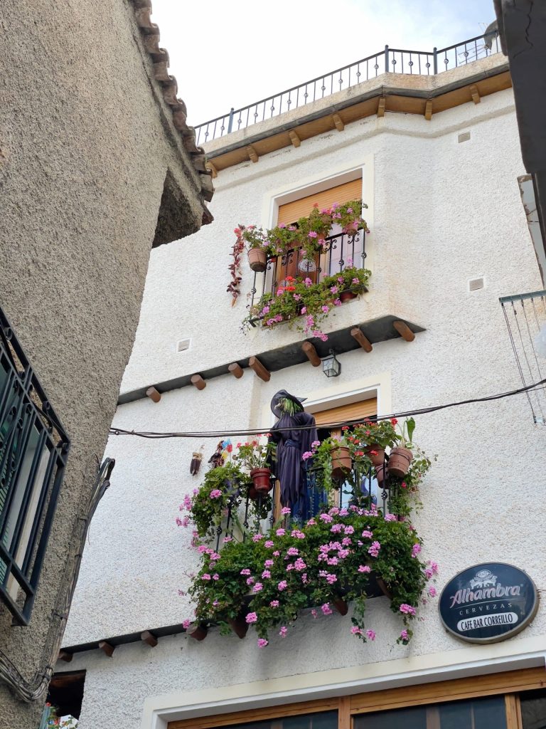 A white building with flower-filled balconies and a statue of a veiled figure among pink and red blooms. A sign reading "Alhambra" hangs above the doorway—an inviting stop for those enjoying day trips from Granada.