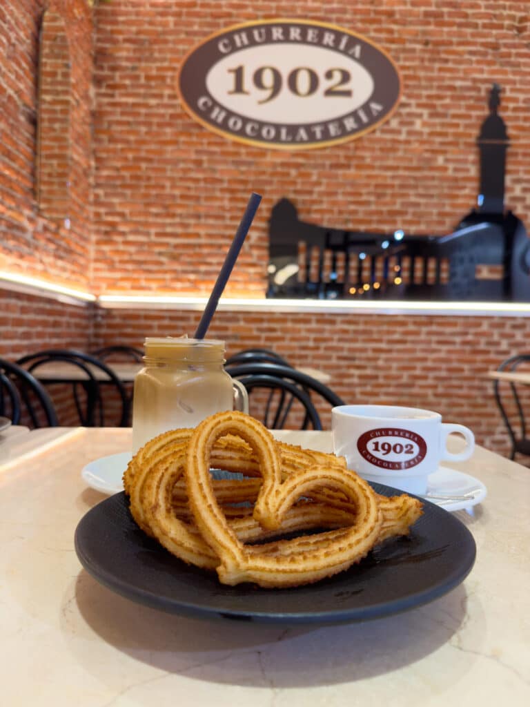 A plate with heart-shaped gluten free churros, an iced coffee with a straw, and a cup of hot chocolate sit on a table in a cafe with brick walls at Chocolatería 1902—a perfect stop for anyone exploring things to do in Madrid.