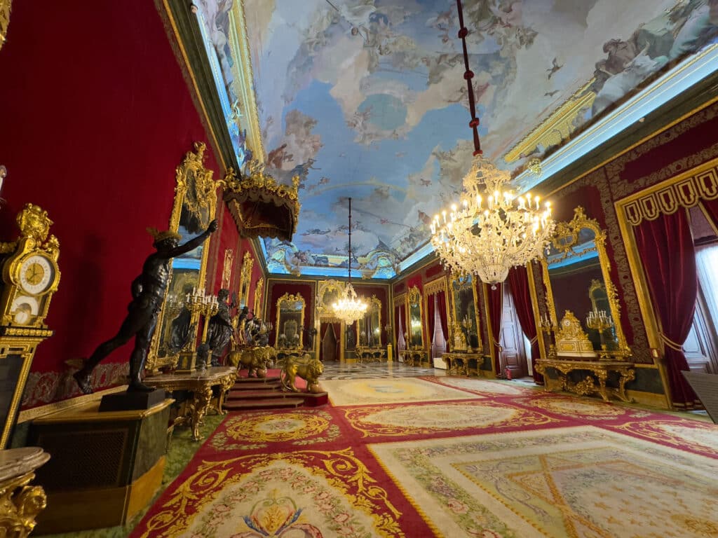 A lavish palace room with red velvet walls, ornate gold-framed mirrors, and grand chandeliers—exploring such opulence is one of the top things to do in Madrid. Patterned carpets cover the floor while elegant furniture lines the walls.