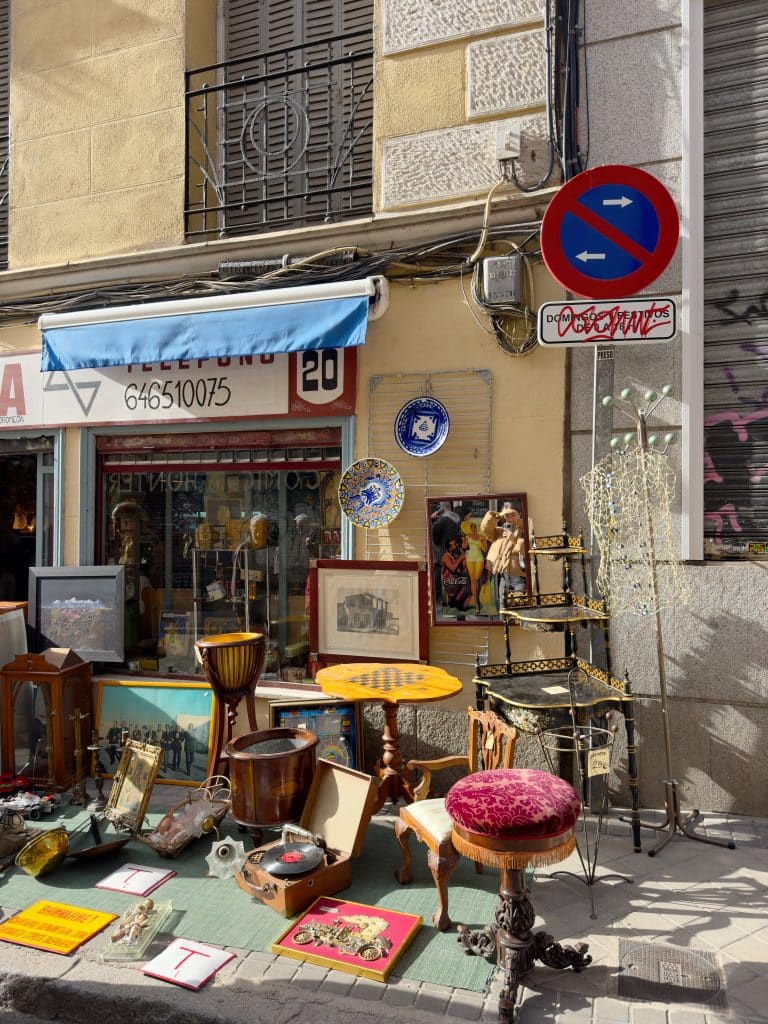 A sidewalk display of vintage items—furniture, framed art, drums, and decorative objects—in front of a shop. A "no parking" sign and a blue building facade are visible above the storefront—perfect for those seeking unique things to do in Madrid.