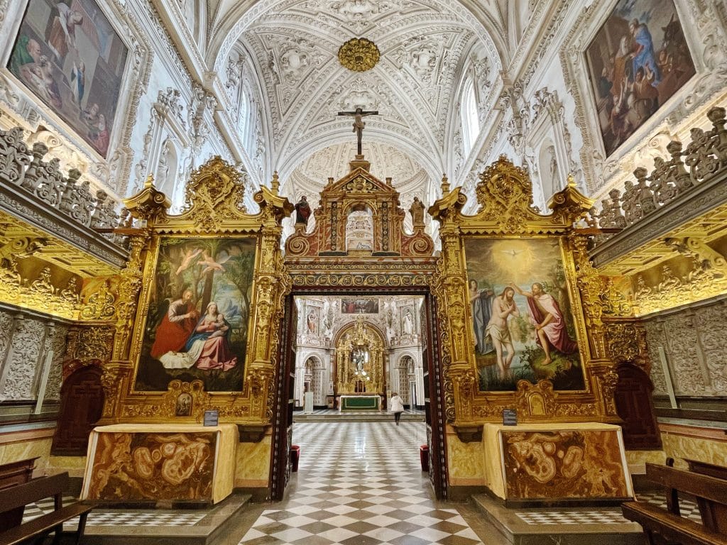 Ornate church interior with intricate white vaulted ceiling, large gold-framed religious paintings, decorative altarpiece, checkered floor, and detailed carvings; this is one of the hidden gems of Granada where colorful murals adorn the walls.