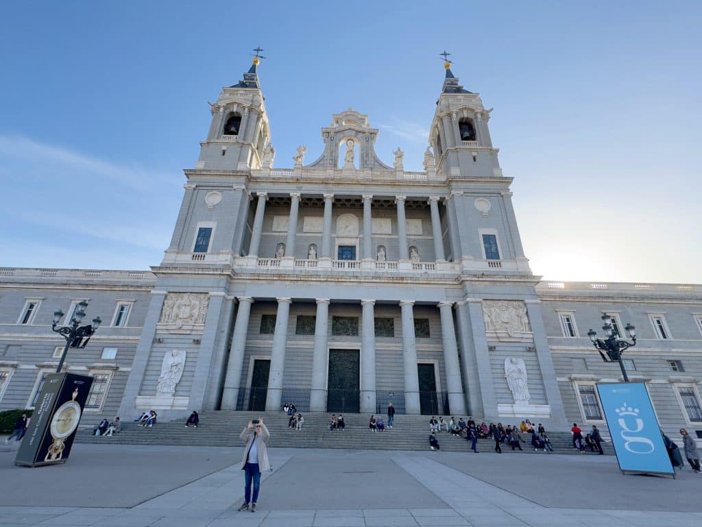 A wide-angle view of a neoclassical cathedral with two tall towers and ornate details. People sit on steps leading up to the entrance. The sky is clear, and banners are displayed on either side of the plaza.