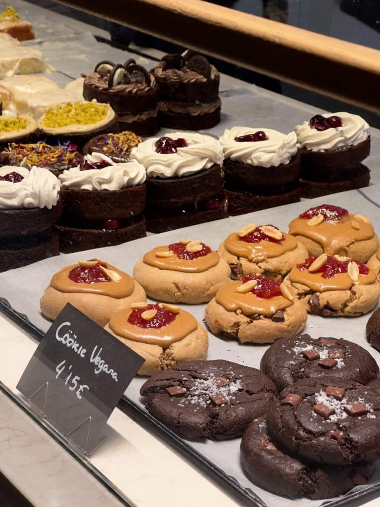 A display of desserts including chocolate cakes topped with cream and cherries, and cookies with jam and almond slices. A sign in the foreground reads "Cookie Vegana 4,15€". The sweets are arranged on a marble counter.