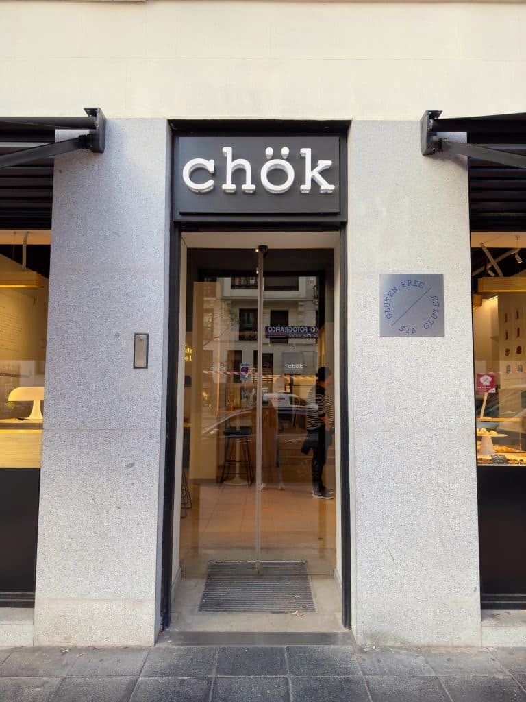 Storefront of "chök," featuring a glass door flanked by stone walls. Inside, a glimpse of the interior shows tables and what appears to be a counter. A sign on the wall indicates gluten-free options are available.