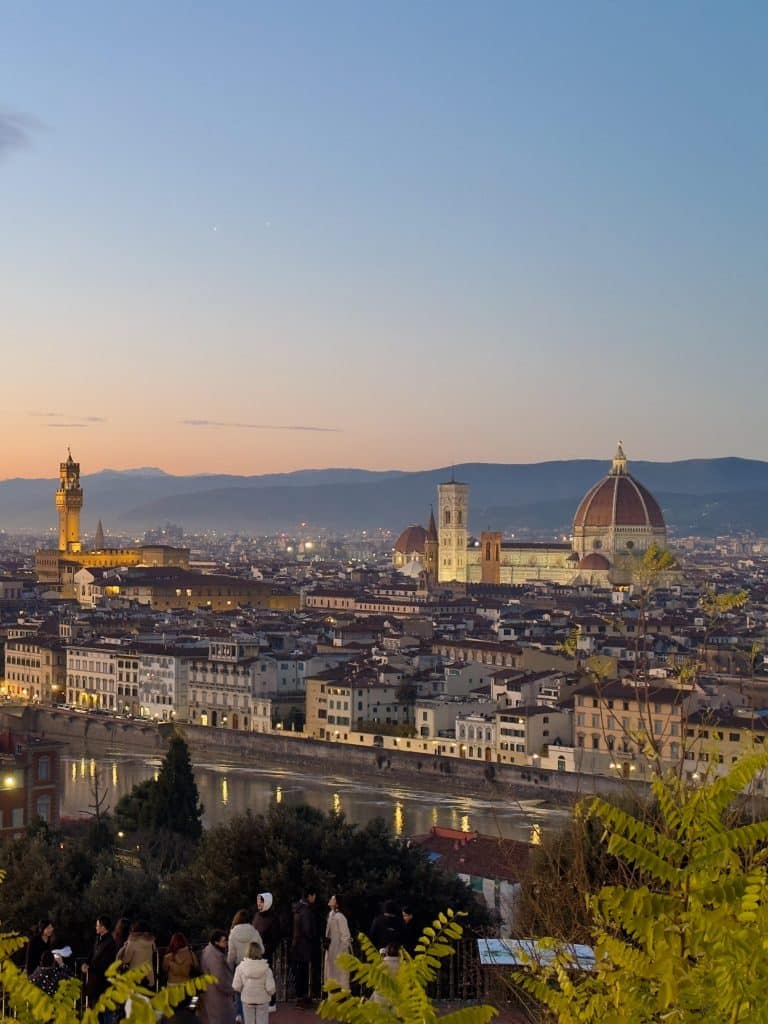 A panoramic view of Florence, Italy at sunset. The iconic Florence Cathedral and Palazzo Vecchio are visible against a colorful sky. The Arno River flows through the city, with hills in the distance and foliage in the foreground.