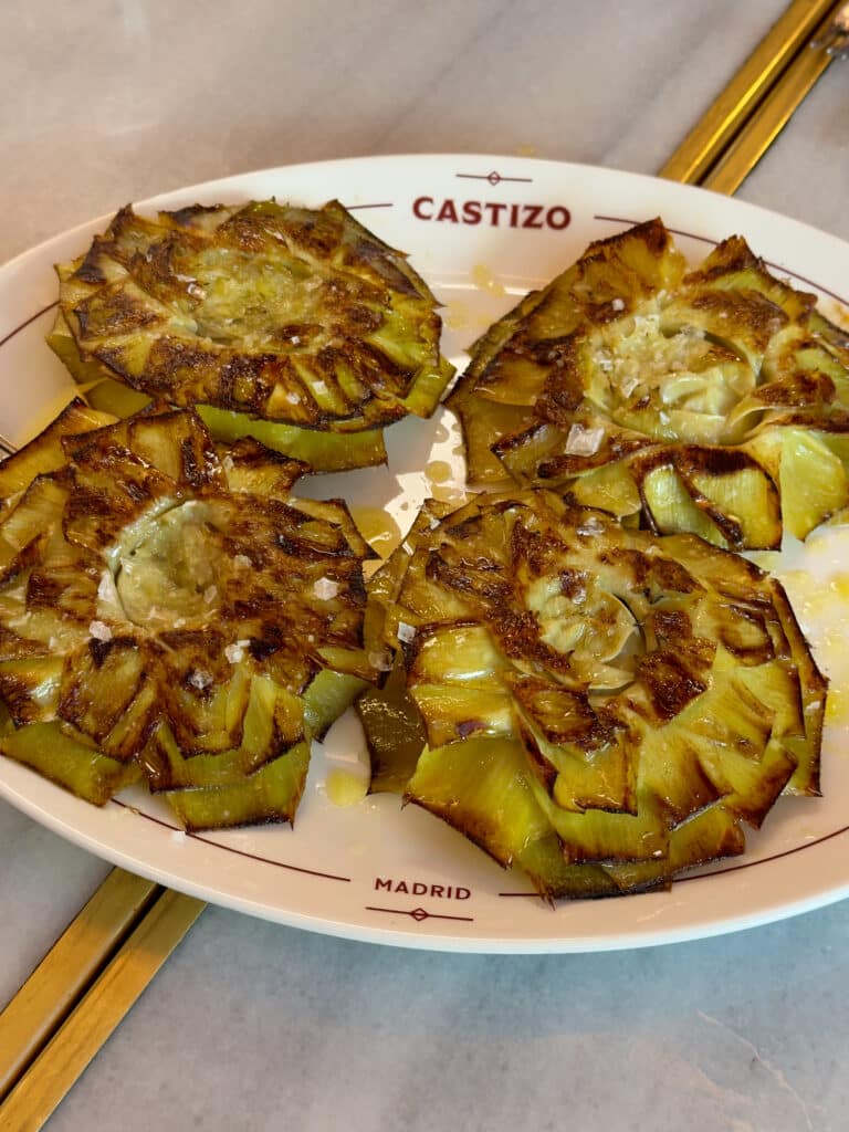 Four crispy, browned roasted artichoke slices arranged on a white oval plate with "CASTIZO" and "MADRID"—a delicious bite to add to your list of things to do in Madrid, drizzled with oil and a sprinkle of salt.