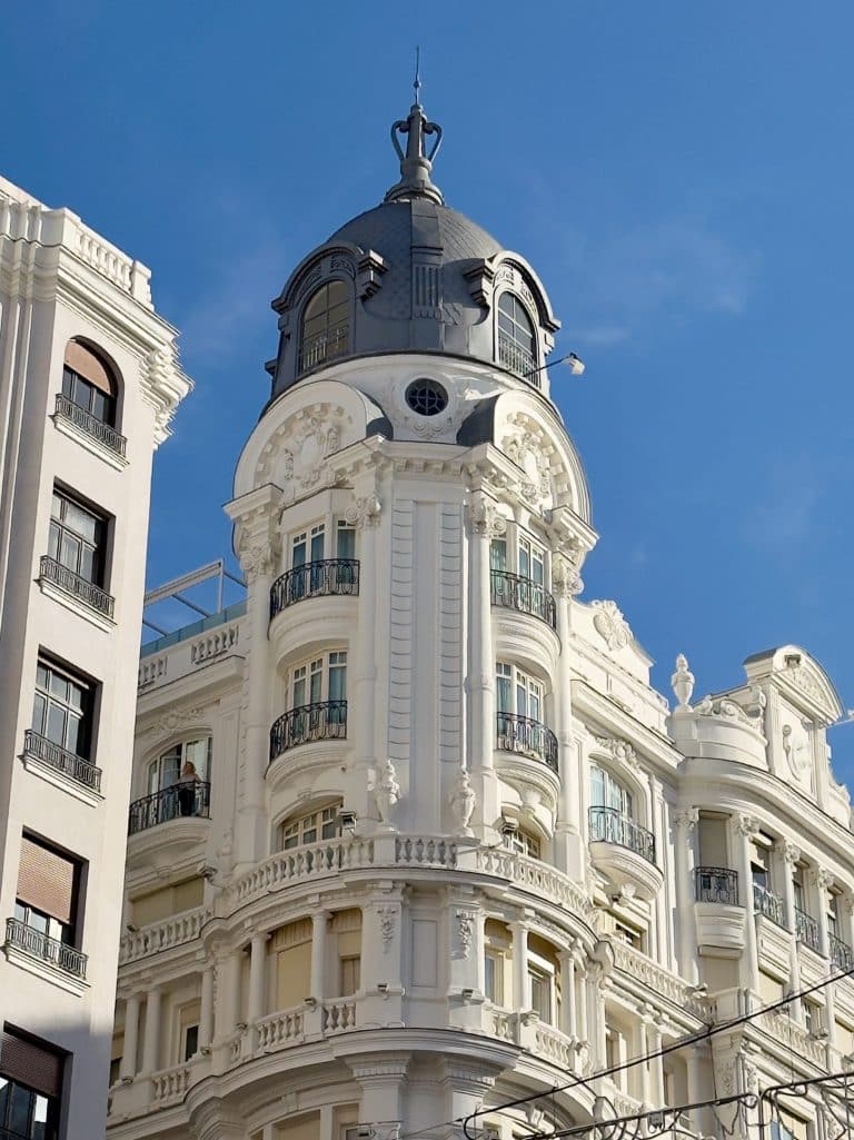 Ornate white building with a tall, rounded tower and intricate architectural details, set against a clear blue sky. The facade features multiple balconies with wrought iron railings and decorative sculptures.