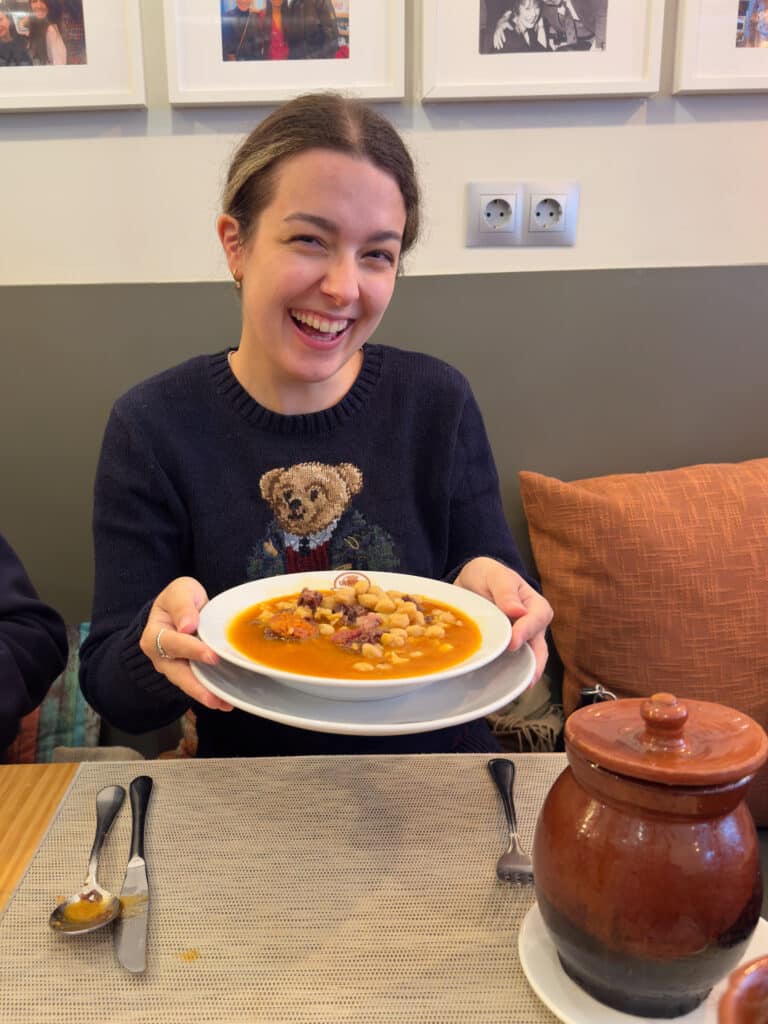 A smiling woman sits at a table holding a plate of Madrid traditional stew, cocido madrileño. She wears a navy sweater with a teddy bear design. A ceramic pot rests on the table, and framed photos hang on the wall behind her.