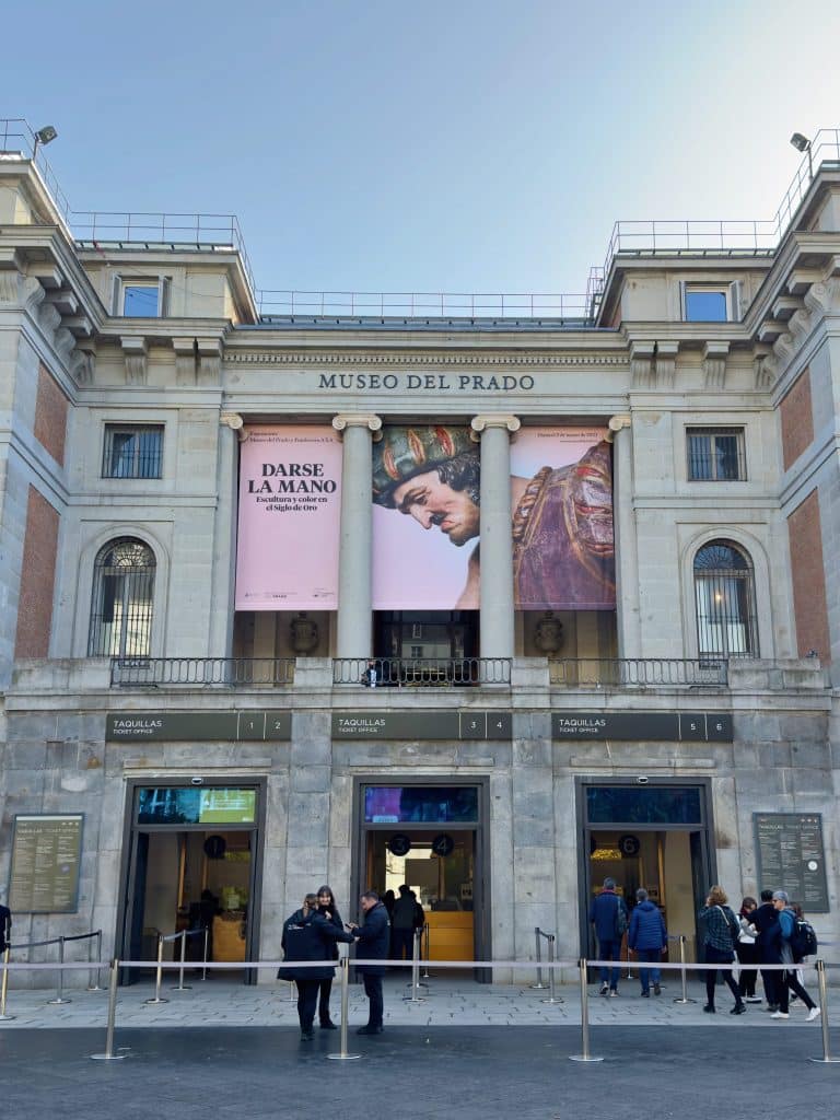 The image shows the front entrance of the Museo del Prado, a renowned art museum in Madrid. The facade features a large advertisement for an exhibition titled "Darse la Mano." Visitors are gathered around the entrance.