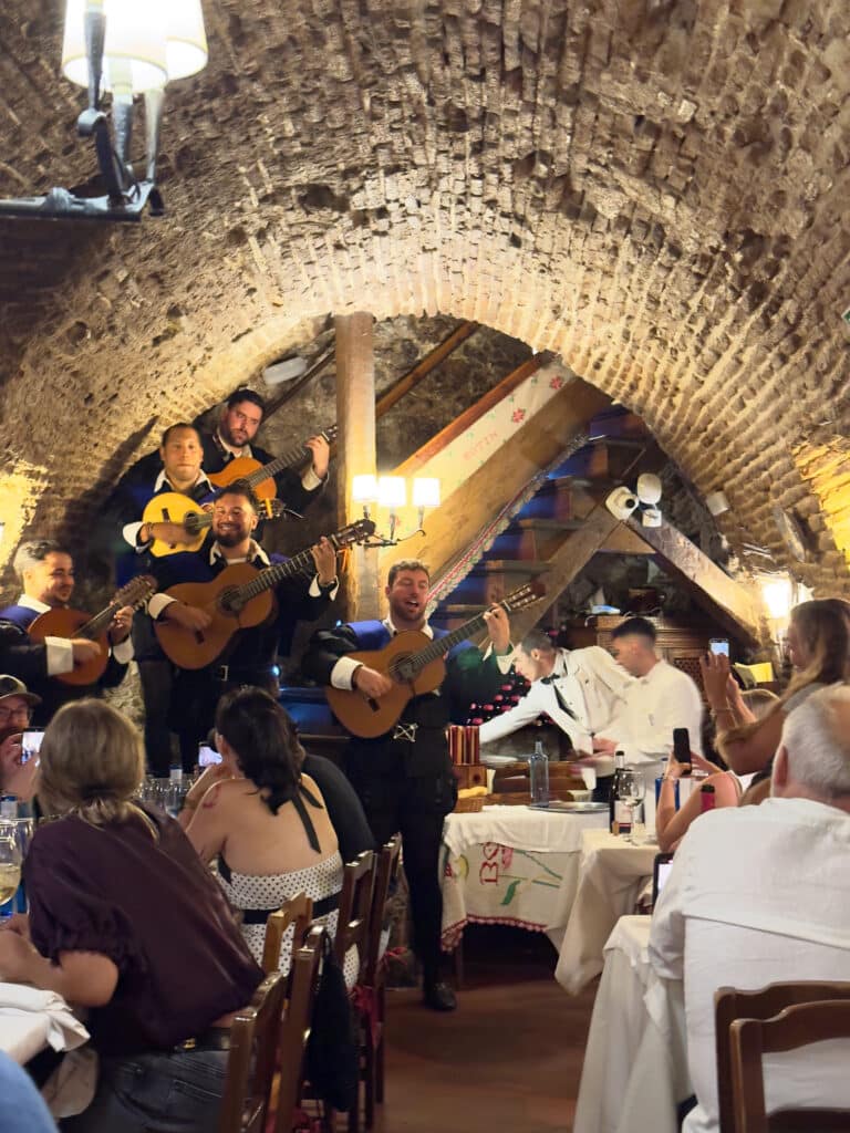 A lively group of musicians, La Tuna, with guitars performs in a rustic, arched brick restaurant, Restaurante Botín, as diners watch, clap, and enjoy their meals at crowded tables under warm lighting—a must-see for those seeking authentic things to do in Madrid.