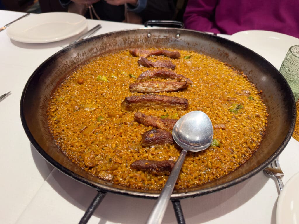 A large shallow pan of rice with pieces of meat arranged in a line on top, served on a white tablecloth with empty plates and utensils around it—a traditional food to try in Madrid. A large serving spoon rests on the edge of the pan.