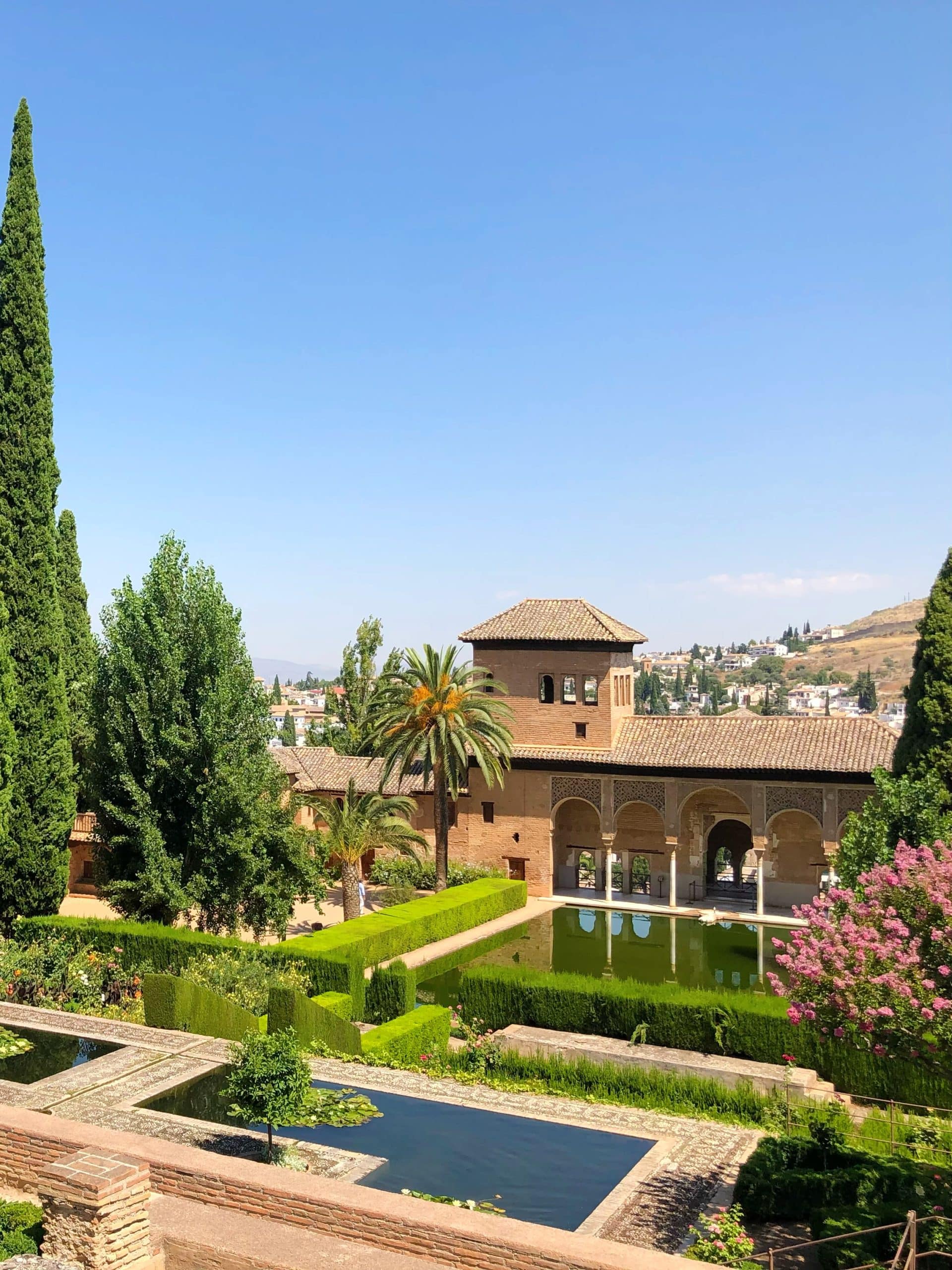 A lush courtyard garden with geometric hedges, tall cypress trees, and a reflecting pool beside a historic stone building—an unforgettable spot if you have just one day in Granada under a clear blue sky.
