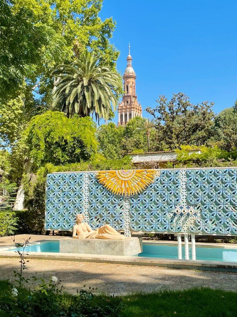 Explore a serene garden scene in Sevilla, Spain, where a reclining statue graces a decorative pool. Behind it, a large patterned mosaic wall boasts a sun motif. Lush greenery and palm trees envelop the area, with an ornate tower rising majestically in the background.