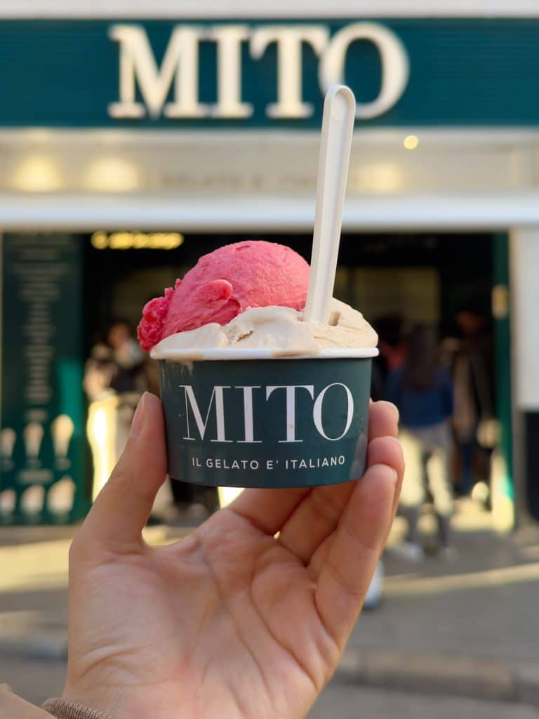 A hand holds a cup of gelato, featuring two delightful scoops—one pink and one beige—with a white spoon. The cup, marked "MITO," captures attention against the blurred backdrop of Sevilla streets, promising gluten-free indulgence in the heart of the city.