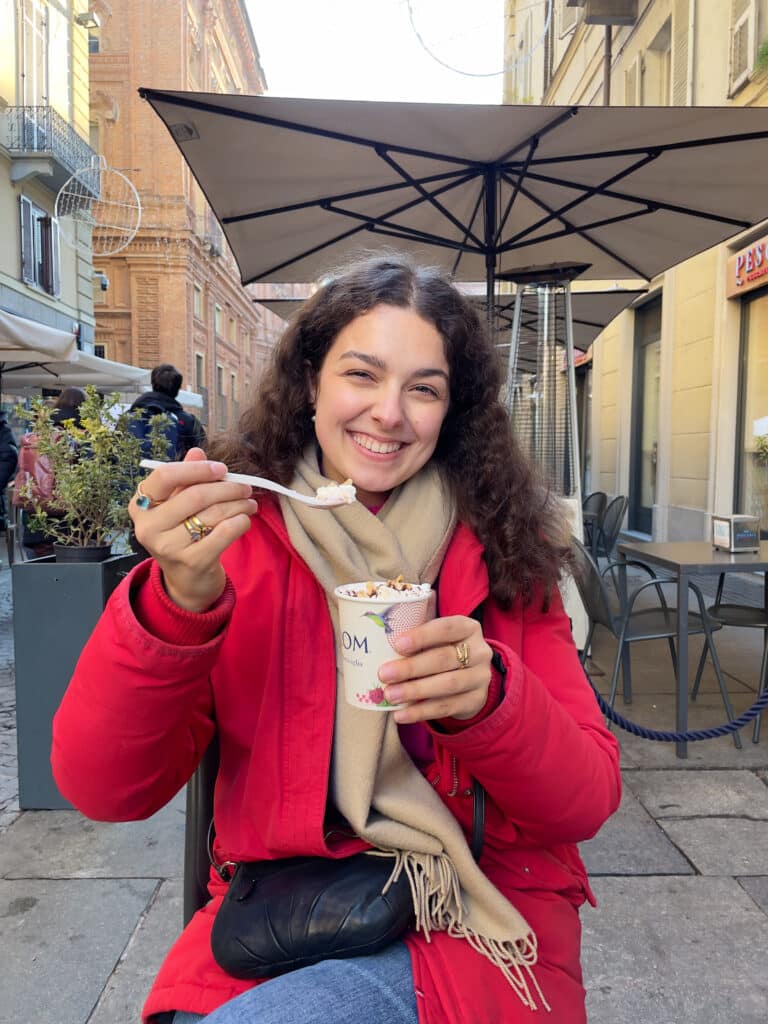 A smiling woman in a red coat and beige scarf sits at an outdoor cafe table, holding a cup of ice cream with sprinkles and eating it with a spoon. She is surrounded by city buildings and umbrellas.