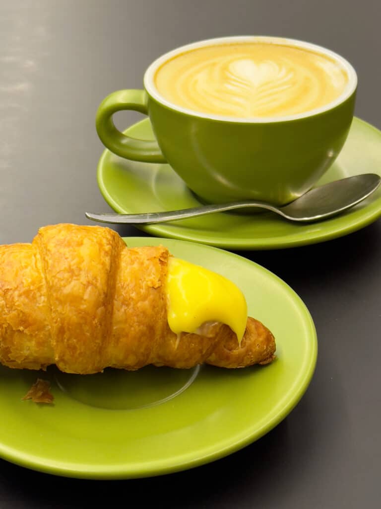 A green cup of latte with latte art and a spoon sits on a saucer next to a croissant filled with yellow custard on a matching green plate, all on a dark surface.