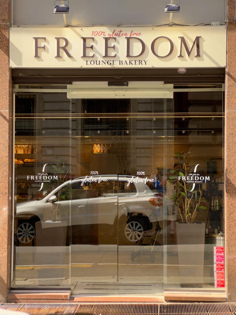Glass storefront of “FREEDOM LOUNGE BAKERY” with “100% gluten free” written above the entrance. Reflections of a parked white car and a building are visible on the glass doors. Indoor plants are seen inside.