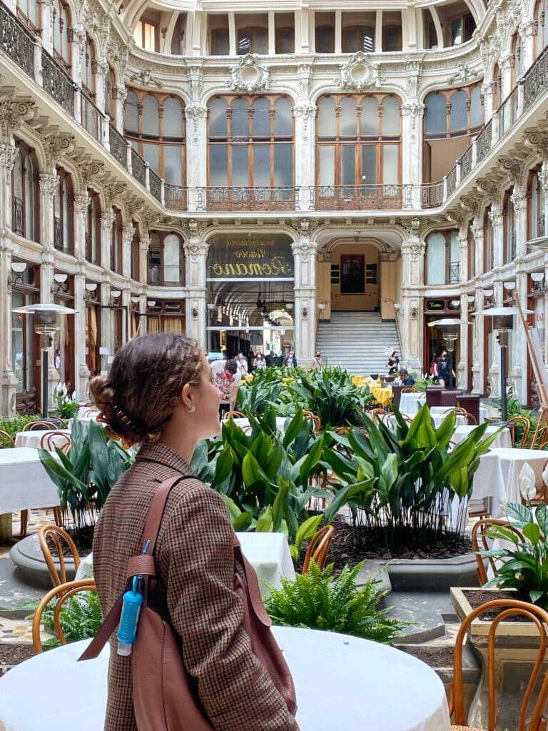 A woman with curly hair and a brown jacket stands in an ornate, indoor courtyard filled with green plants and tables, looking toward a grand staircase and large windows—just one of many reasons to visit Turin.