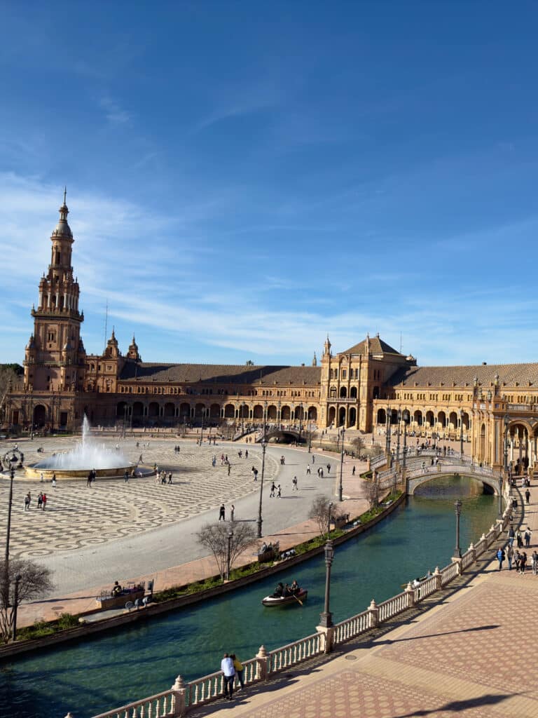 Experience the charm of Plaza de España in Seville, Spain, on your one day in Seville. Marvel at its semicircular Renaissance Revival architecture, a central fountain, and a picturesque canal with small bridges, all under a clear blue sky as visitors leisurely stroll by.