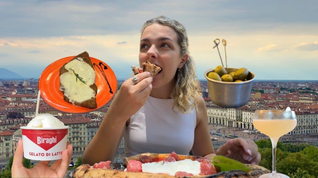 A woman eats pizza at an outdoor table overlooking a cityscape. Around her are a slice of bread with melted cheese, a bowl of green olives, a cup of gelato, and a cocktail. The sky is cloudy in the background.