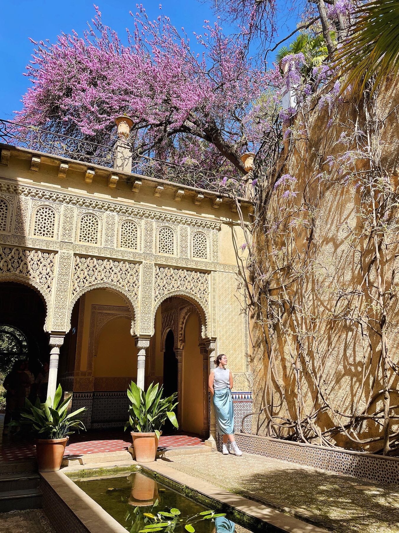 A woman stands beside a long wall covered in vines and purple blossoms in a sunlit courtyard—one of the hidden gems of Granada—with ornate arches, potted plants, and a small reflecting pool under a bright blue sky.