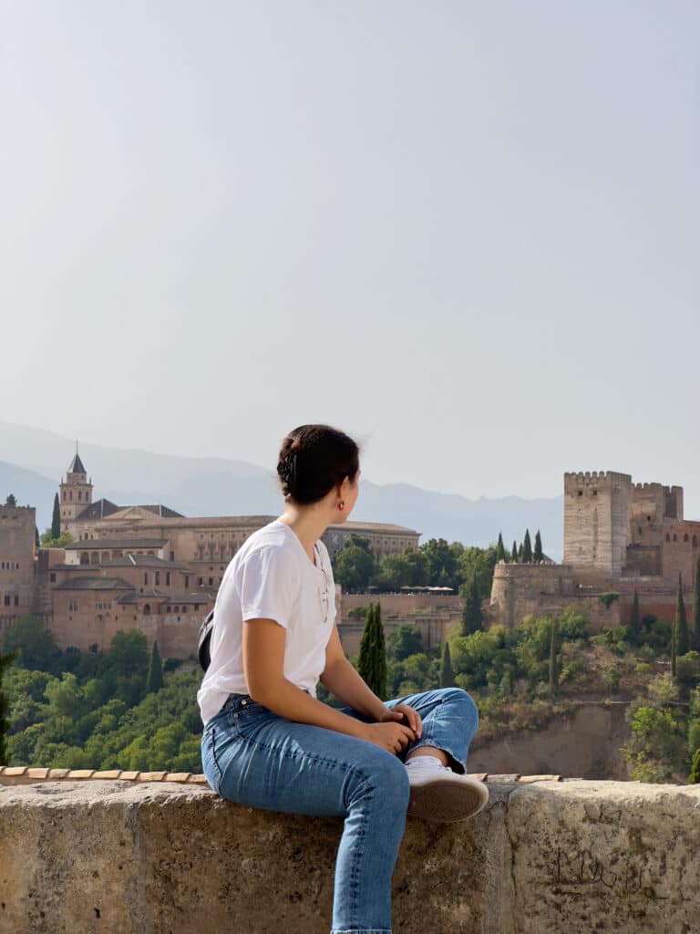 A person in a white shirt and jeans sits on a stone ledge, looking toward a large historic fortress surrounded by trees and mountains under a clear sky.