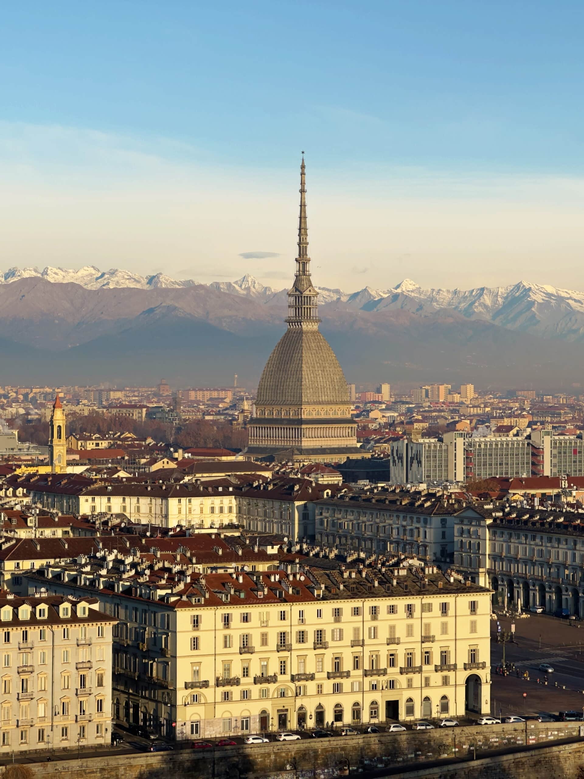 A panoramic view of Turin, Italy, from Monte dei Cappuccini, featuring the iconic Mole Antonelliana at the center, historic buildings all around, and snow-capped mountains in the background—scenery like this is one of many reasons to visit Turin.