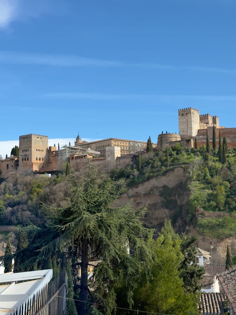 Views of the Alhambra, a historic palace and fortress complex, crowns a tree-covered hill under a clear blue sky from Carmen El Agua restaurant -a must-eat at when in Granada.