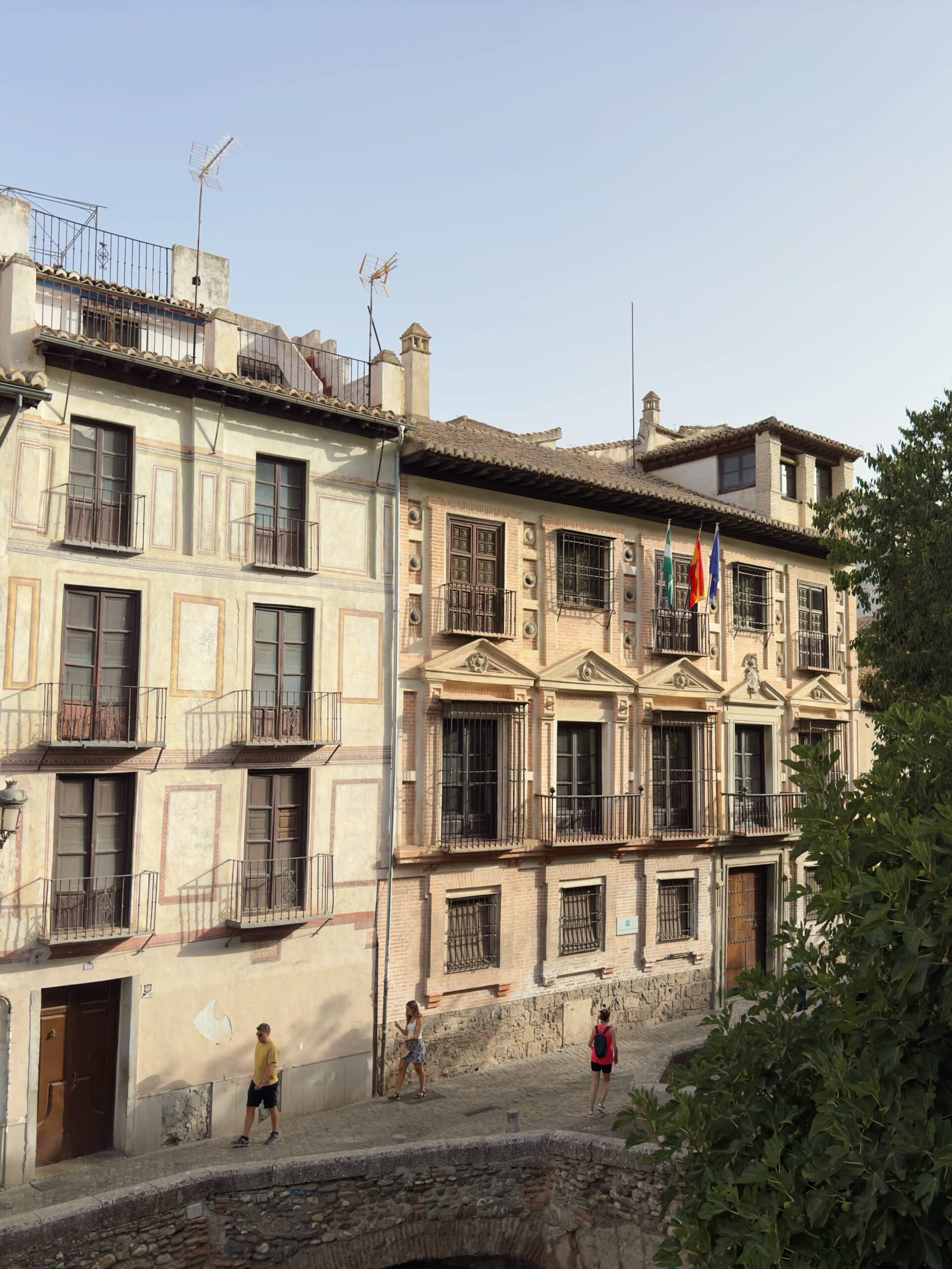 Street view of old European buildings with balconies and Spanish flags, trees on the right, and two people walking on a stone path below—a charming scene perfect for any two days in Granada. The sky is clear and bright.