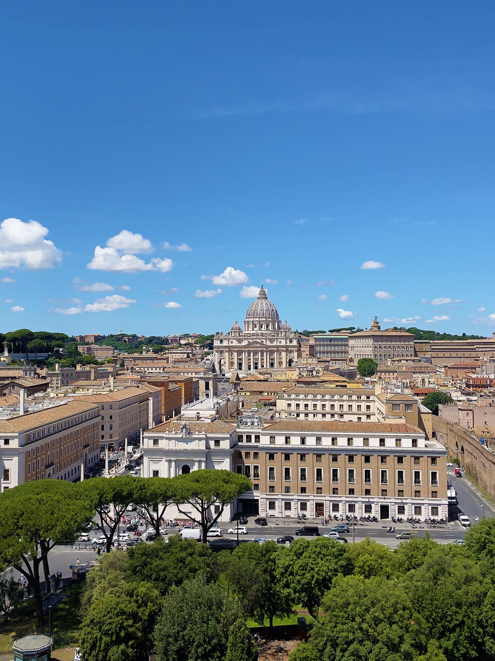 A panoramic view of Vatican City with St. Peter’s Basilica at the center, a highlight for any 2 days in Rome itinerary, surrounded by historic buildings, trees in the foreground, and a bright blue sky with scattered clouds.