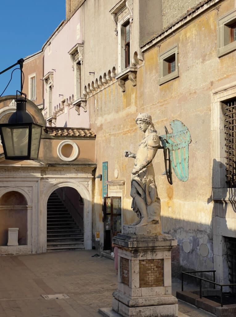 A stone statue of a man stands on a pedestal in a sunlit courtyard with weathered yellow and pink walls, arched doorways, windows, and a hanging streetlamp—a charming scene to include in your 2 days in Rome itinerary. Stairs are visible in the background.