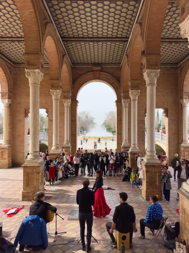 A group of musicians and a flamenco dancer perform under an ornate arched pavilion as a crowd watches. Sunlight streams through, illuminating the scene, with a fountain and trees visible in the background.