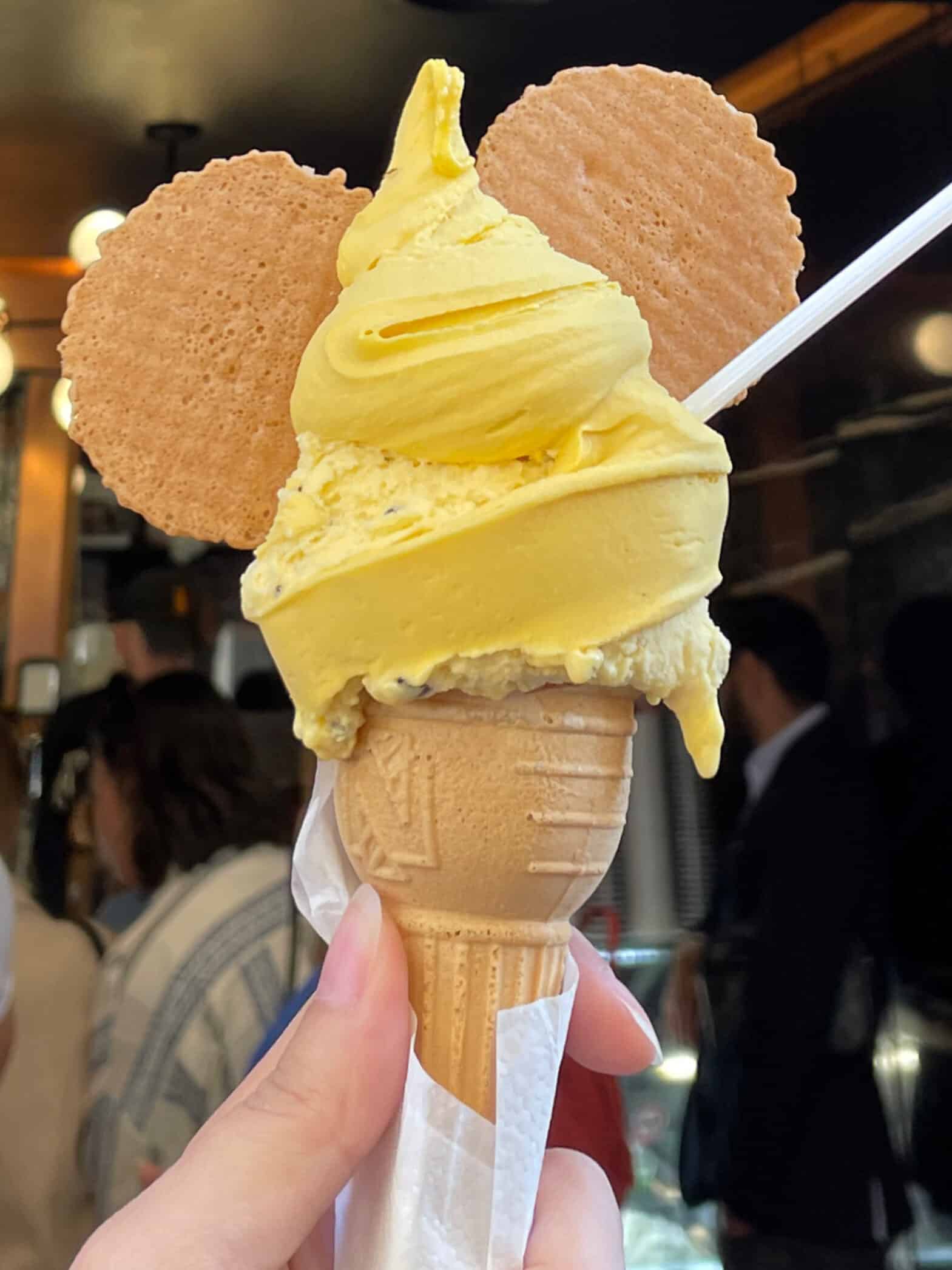 A hand holds a gluten free gelato cone with yellow gelato and two round cookies resembling Mickey Mouse ears, with a small spoon inserted—a sweet treat you might discover on your 2 days in Rome itinerary. People and lights blur softly in the background.