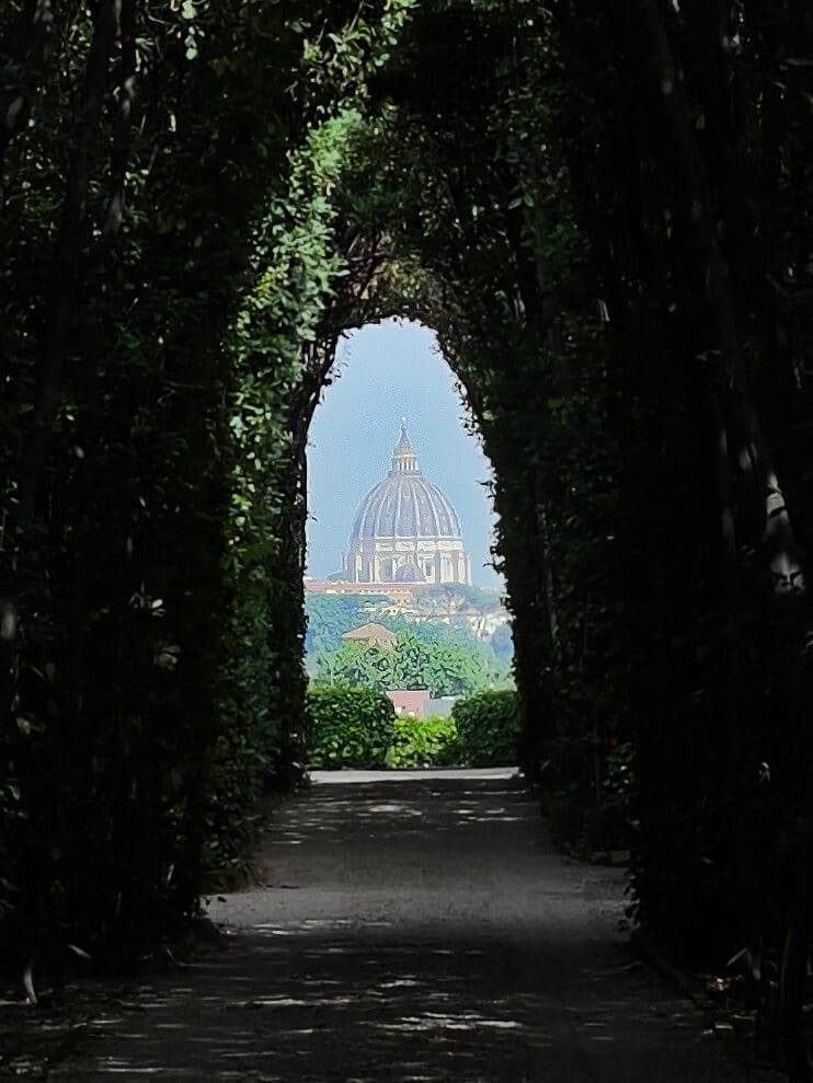 A tree-lined tunnel frames a distant view of the dome of St. Peter’s Basilica in Vatican City—an iconic scene to discover during your 3 days in Rome itinerary, with sunlight illuminating both lush greenery and the famous dome beyond.