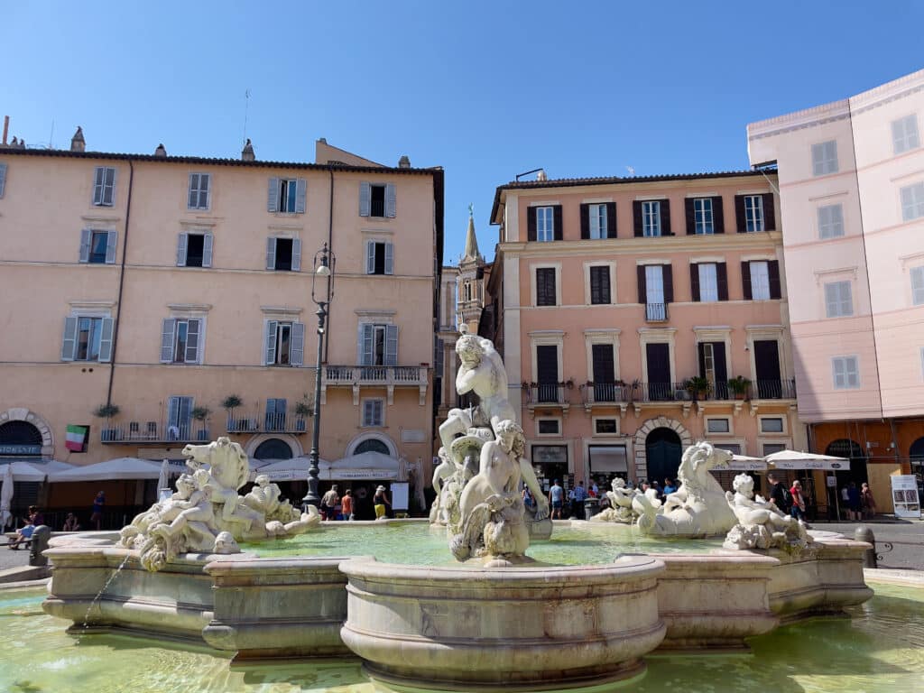 A highlight of any 2 days in Rome itinerary, a large stone fountain with sculpted figures and water at its base stands in a sunny plaza, Piazza Navona, surrounded by pastel-colored buildings with windows and awnings under a clear blue sky.