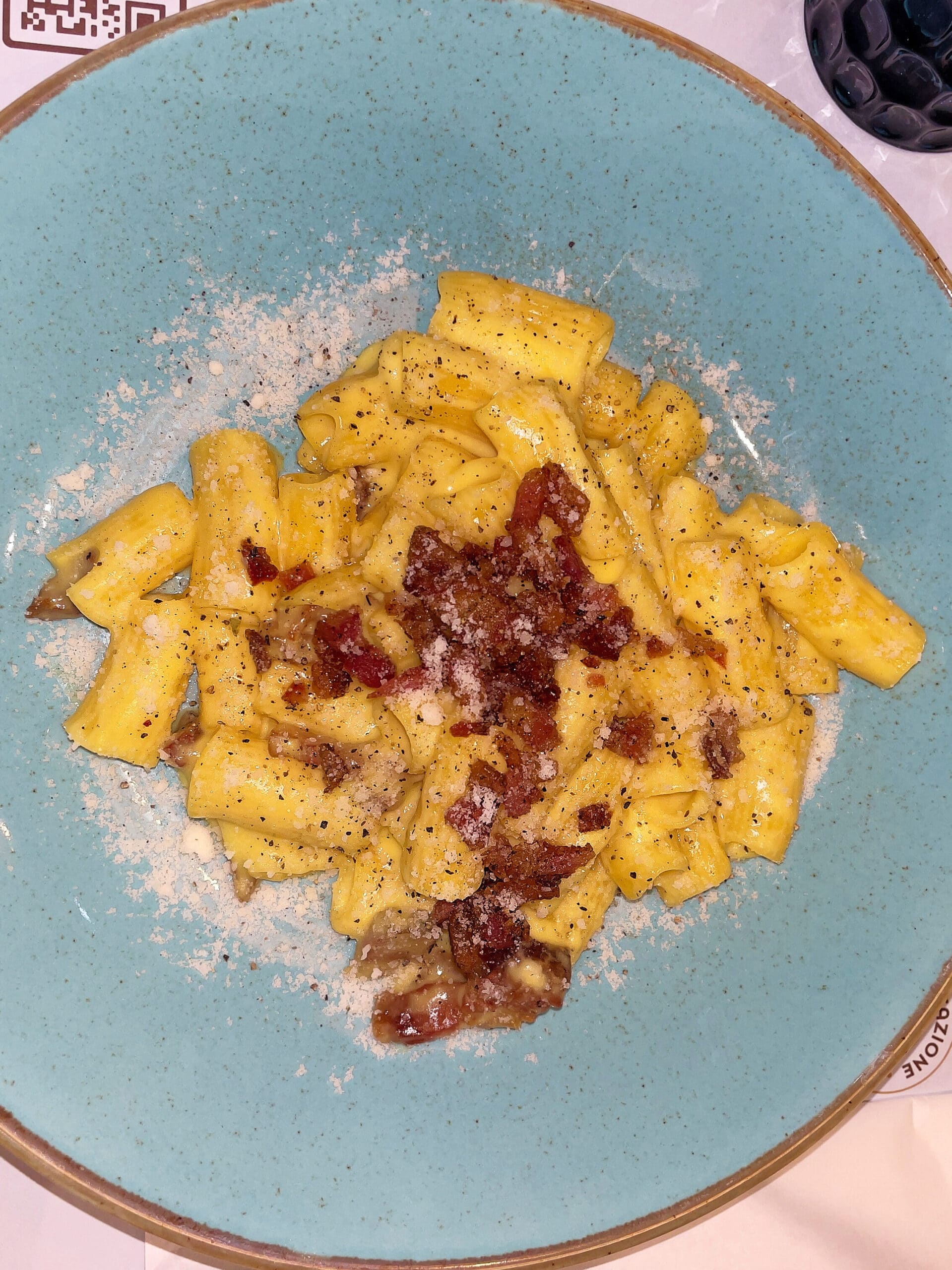 A plate of gluten free rigatoni pasta in a creamy Carbonara sauce, topped with crispy bacon pieces, grated cheese, and black pepper, served on a blue ceramic dish—perfect inspiration for your 2 days in Rome itinerary.