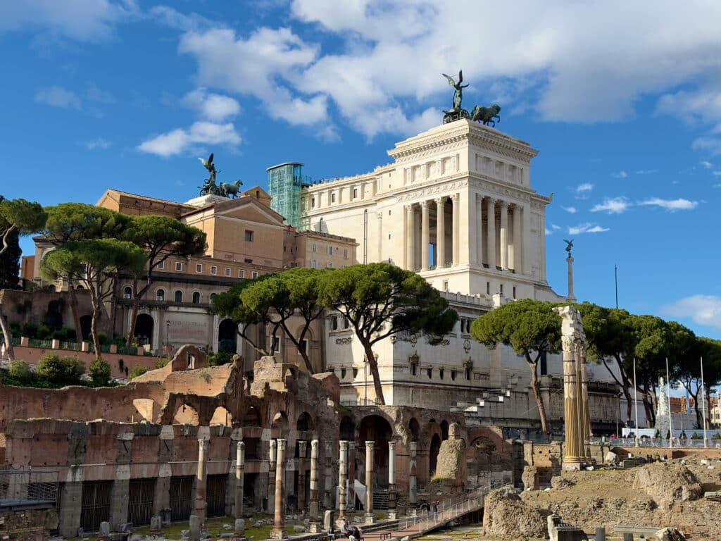 Ancient Roman ruins with stone columns and arches in the foreground, set against the white Altare della Patria monument—an iconic sight you’ll encounter on your 2 days in Rome itinerary beneath a vibrant blue sky.