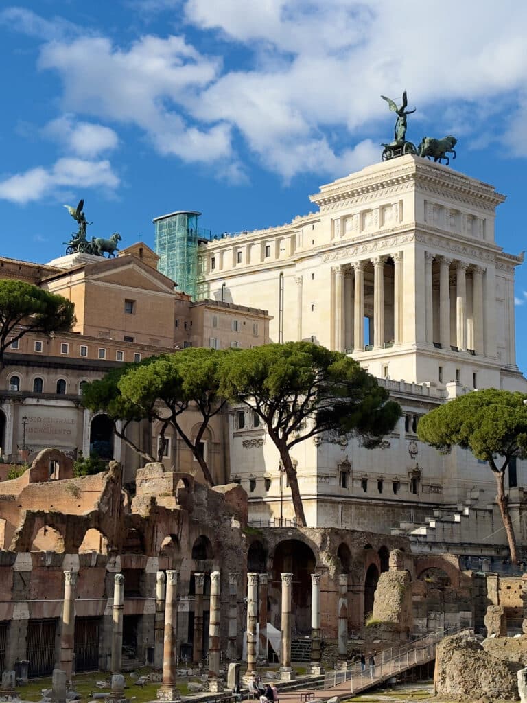 Ancient Roman ruins with stone columns and arches in the foreground, set against the white Altare della Patria monument—an iconic sight you’ll encounter on your 2 days in Rome itinerary beneath a vibrant blue sky.