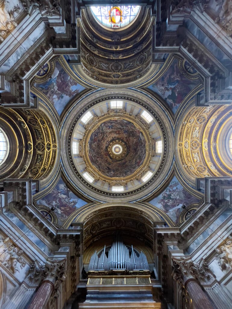 Upward view of an ornate church dome with intricate gold detailing, frescoes, arched windows, and a grand pipe organ—an architectural highlight perfect for your 3 days in Rome itinerary. The richly decorated interior is beautifully symmetrical.
