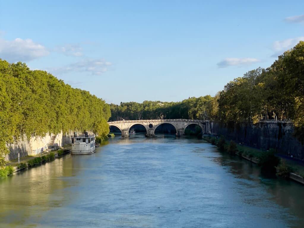 A calm river flows under a stone arch bridge, flanked by lush green trees and a white docked boat on the left side beneath a clear blue sky, evoking tranquil moments you might find on a 2 days in Rome itinerary.