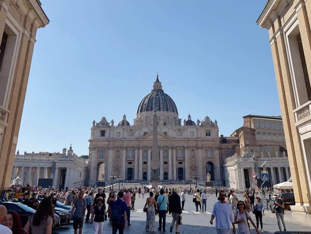 Crowds of people walk in St. Peter’s Square in Vatican City, a must-see stop during 3 days in Rome, facing St. Peter’s Basilica with its large dome and central obelisk under a clear blue sky. Historical columns and buildings frame the scene.