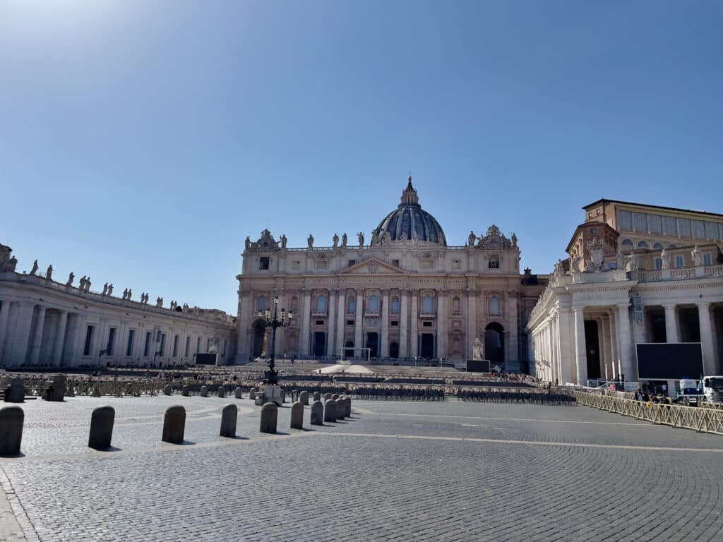 St. Peter’s Basilica in Vatican City under a clear blue sky, featured in many 2 days in Rome itinerary guides, with the square mostly empty and sunlight casting shadows on the cobblestone pavement as statues line the colonnades.