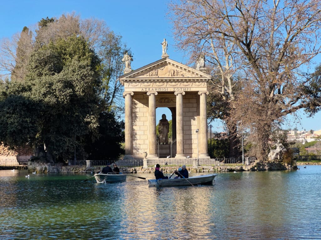 People row boats on a lake in front of an ancient Roman-style temple, a hidden gem you might discover during your 2 days in Rome itinerary, surrounded by trees under a clear blue sky.
