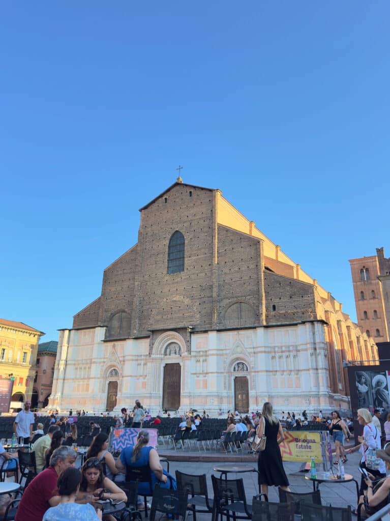 People sit at outdoor tables in a busy square in front of the unfinished facade of the Basilica of San Petronio in Bologna, Italy, under a clear blue sky.