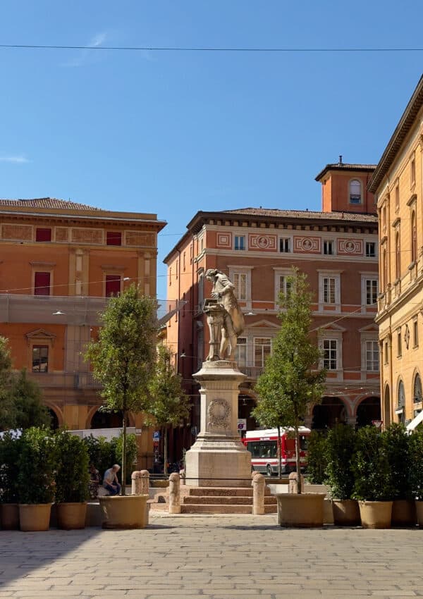 A stone statue of a standing figure on a pedestal is in the center of a sunny plaza, surrounded by potted trees and historic buildings with arched windows and warm-colored facades.
