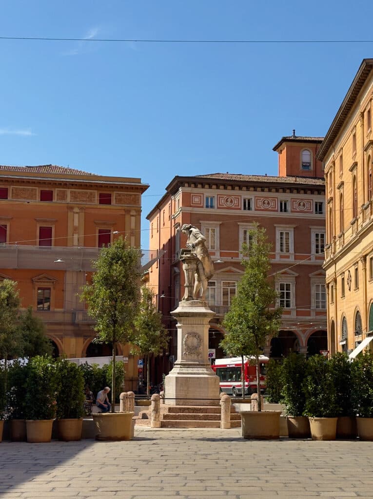 A stone statue of a standing figure on a pedestal is in the center of a sunny plaza, Piazza Galvani in Bologna, Italy, surrounded by potted trees and historic buildings with arched windows and warm-colored facades.