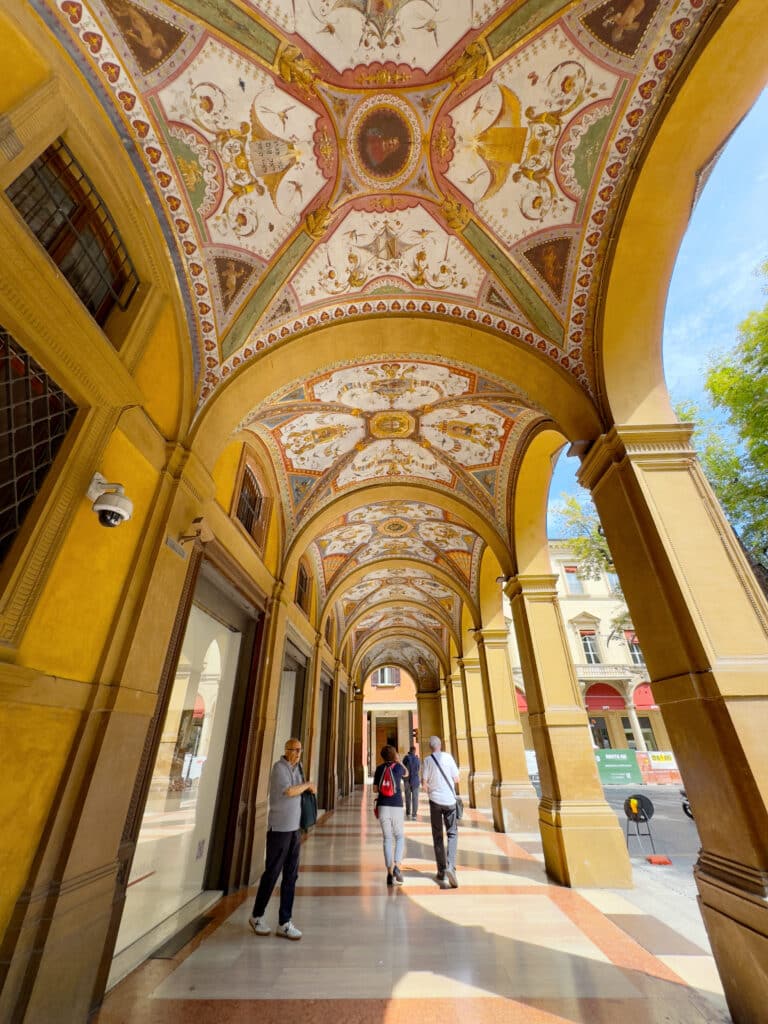 People walk under a historic portico of Bologna with ornate, colorful frescoes on the arched ceiling. Yellow columns and sunlight create a warm, inviting atmosphere in this outdoor gallery.