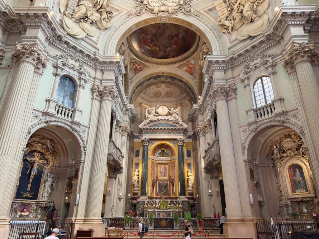 Interior of an ornate cathedral, Santa Maria della Vita, with tall columns, intricate sculptures, religious paintings, and a detailed domed ceiling. Several people are seen admiring the richly decorated altar at the center.